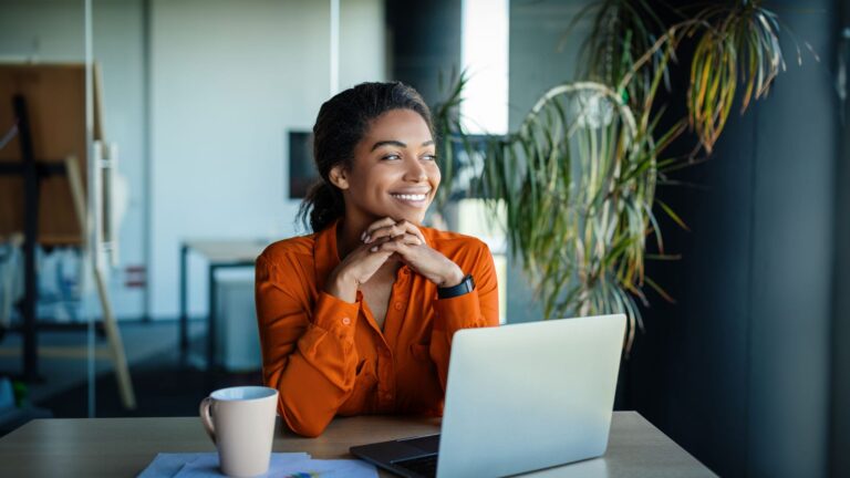 A professional woman in an orange blouse smiles confidently while looking away from her laptop in a bright office space, contemplating strategies like 'how to make your website sell for you.'