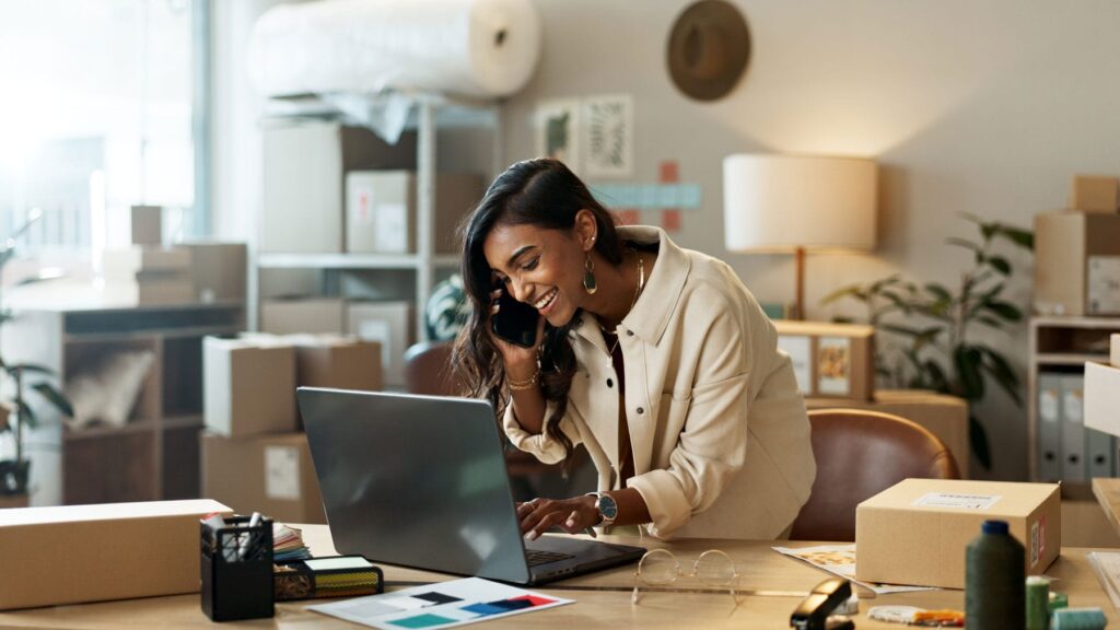 Entrepreneur managing her e-commerce website from a vibrant workspace, conversing on the phone while interacting with a laptop.